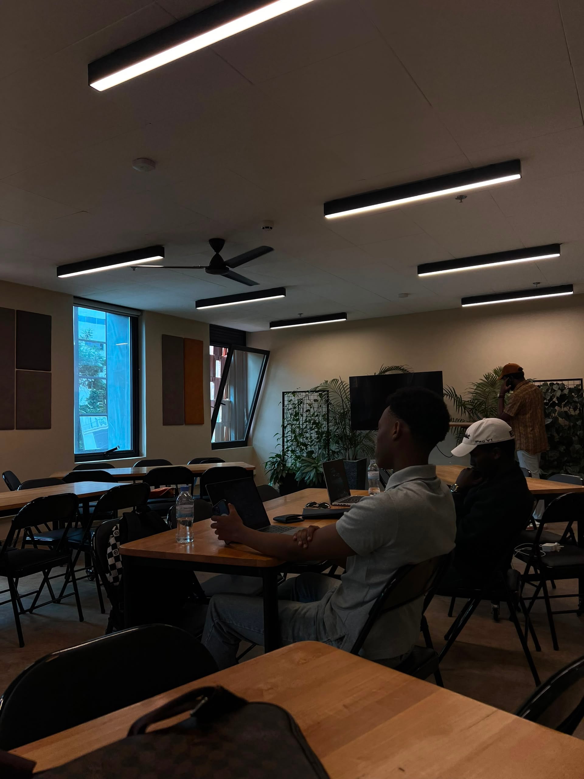 People working on laptops at wooden tables in a bright coworking room with plants and linear ceiling lights