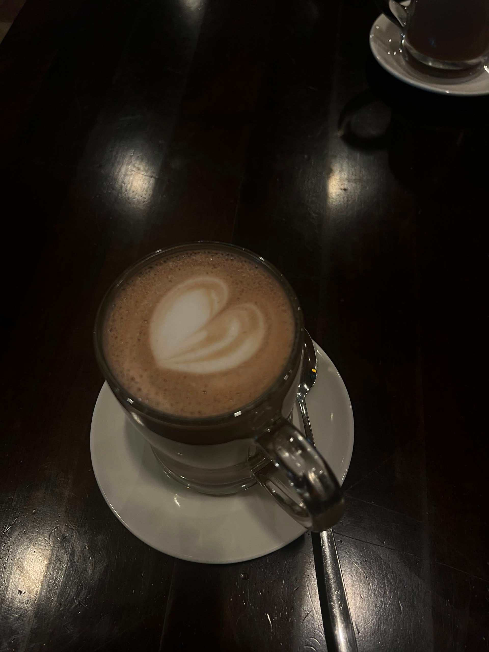 Clear glass mug of latte with heart foam art on a white saucer over a dark wood table