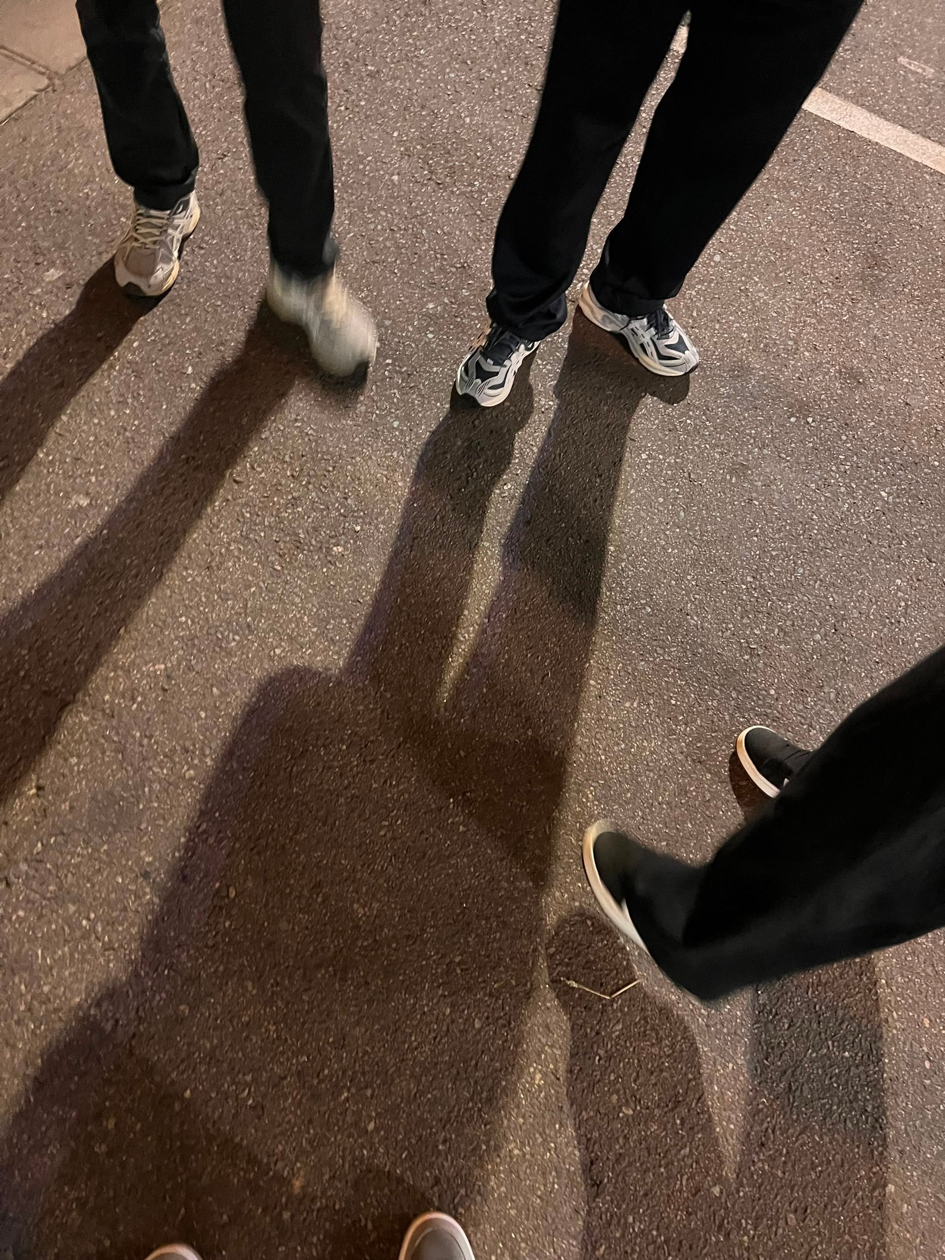 Looking down at several pairs of sneakers and long shadows on asphalt at night
