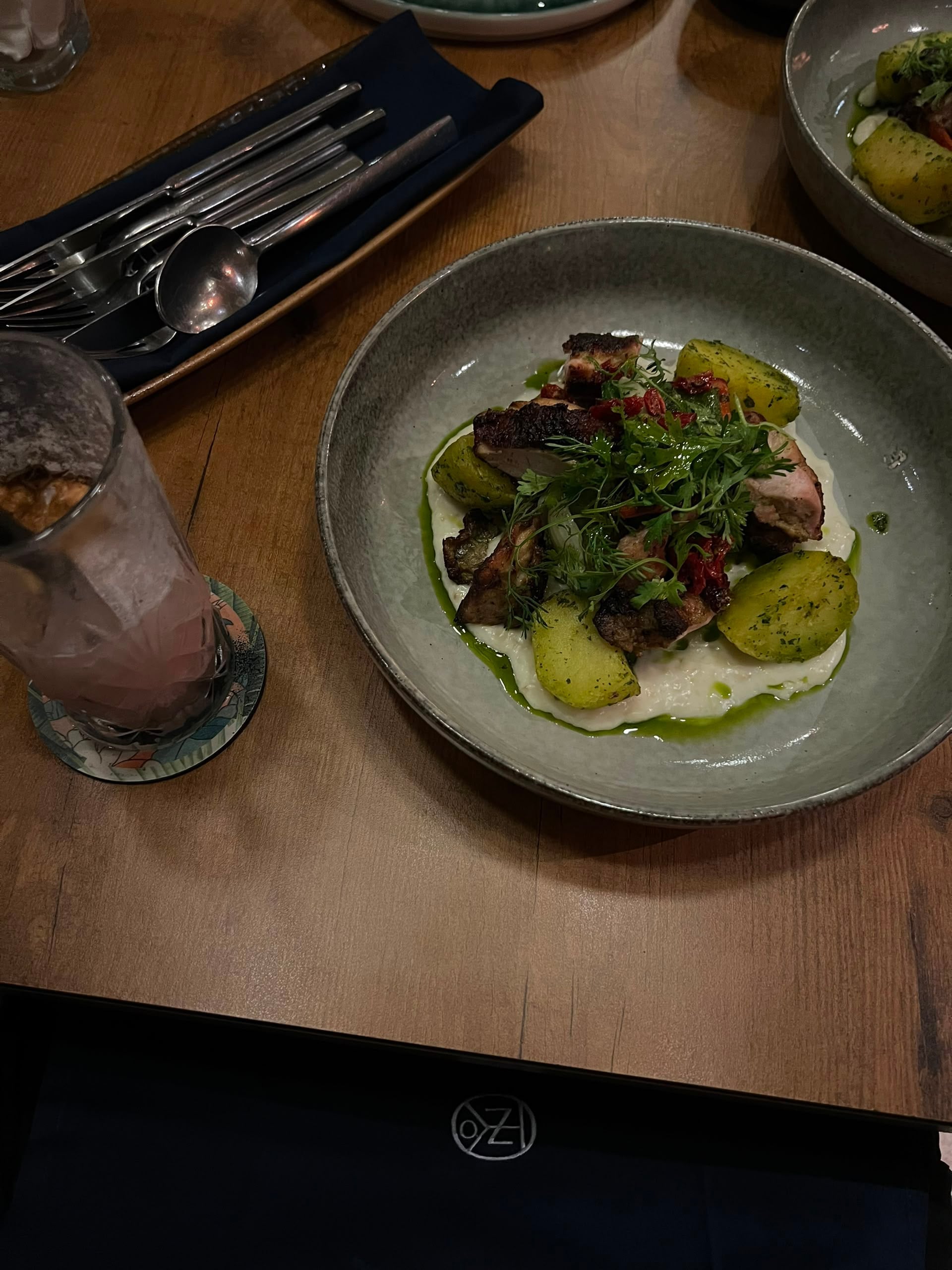 Overhead view of roasted meat and potatoes over white puree with green herb oil, next to a pink drink