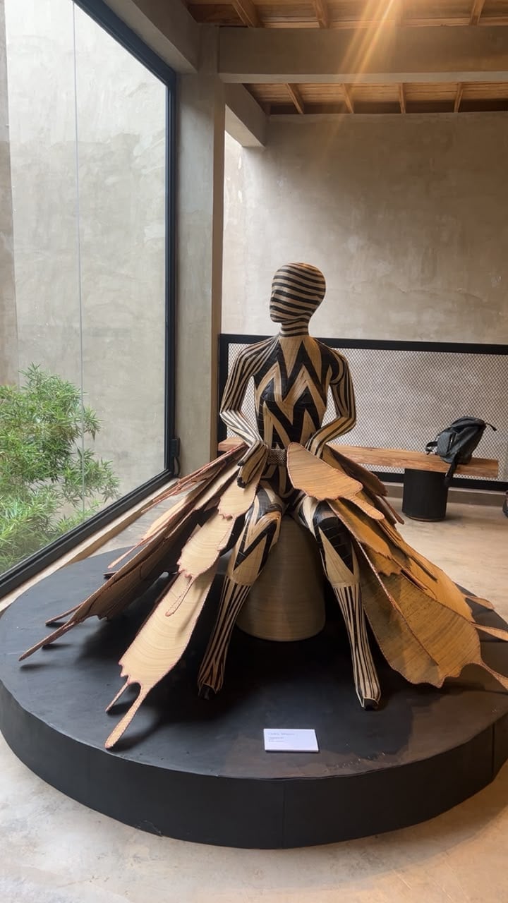 Life-sized striped figurative sculpture with a leaf-panel skirt on a black platform in a concrete gallery